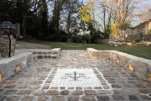 Image montrant une mosaîque en pierres, intégrant une fleur de lys noir en basalte sur un fond de pierres calcaire beige dans une allée en pavés.