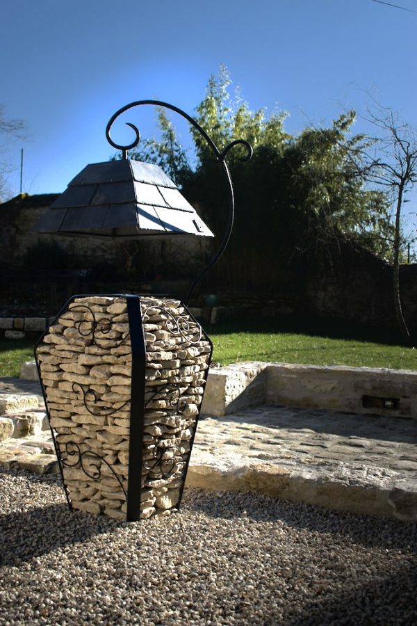 Image montrant une lampe extérieur en pierres et acier avec un abat jour en ardoises dans un jardin avec une allée en pavés.