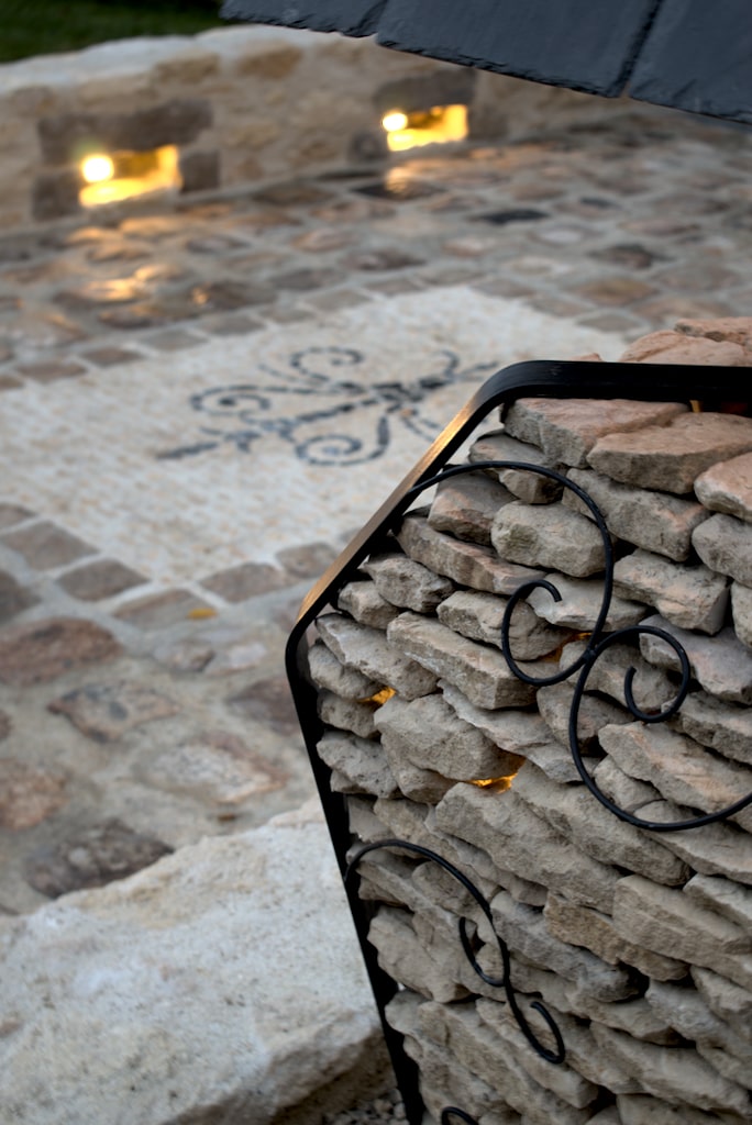Image montrant une partie d'un luminaire extérieur en pierres et aciers, sur une sublime allée en pavés entouré de murs de pierre intégrant des éclairages.