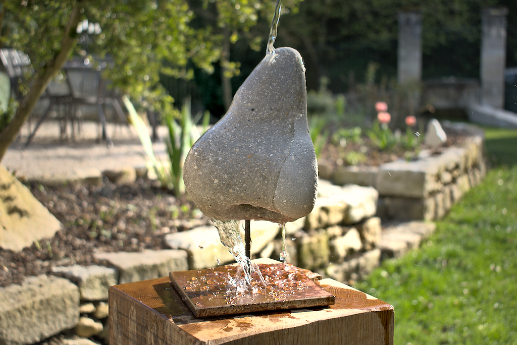 sculpture d'un nez en béton gris sur un socle en acier, arrosée par de l'eau