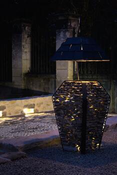 Image montrant une lampe extérieur en pierres et acier avec un abat jour en ardoises dans un jardin avec une allée en pavés.